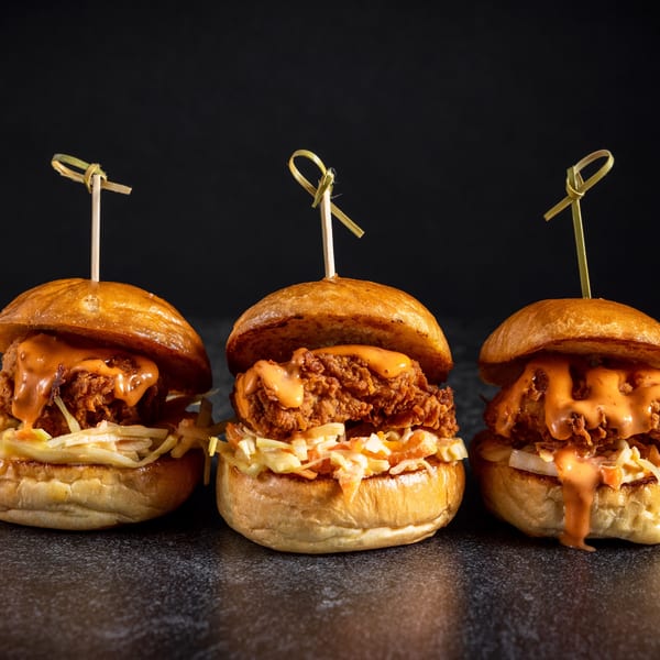 Three Sin City Burger sliders with fried chicken, coleslaw, and orange sauce on dark background.